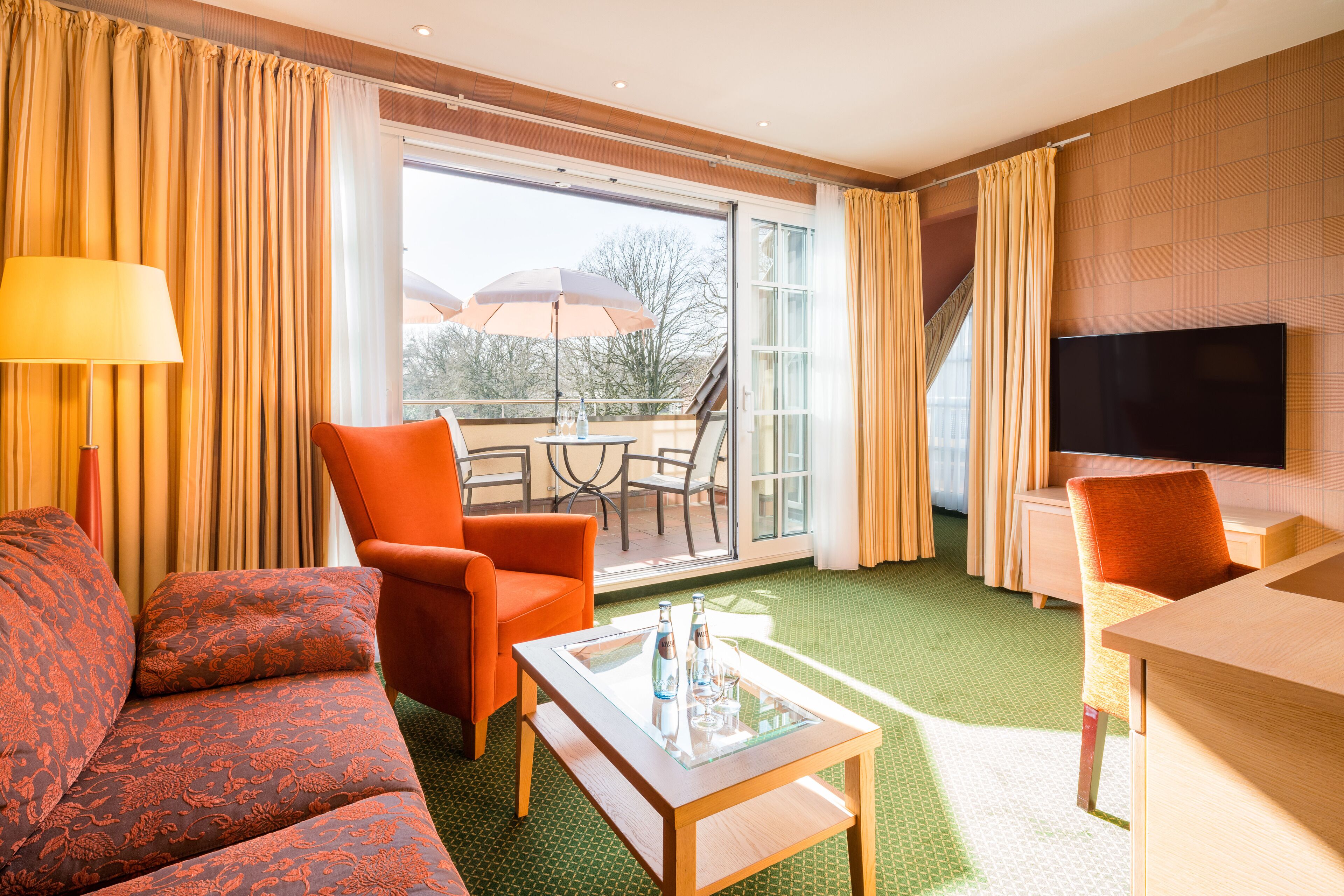 A hotel room with a sofa, two armchairs, a glass-top coffee table, a TV, and a balcony with a table and chairs.