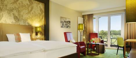 A hotel room with a large bed, a desk, two chairs, a small table with a plant, and a balcony with a view.