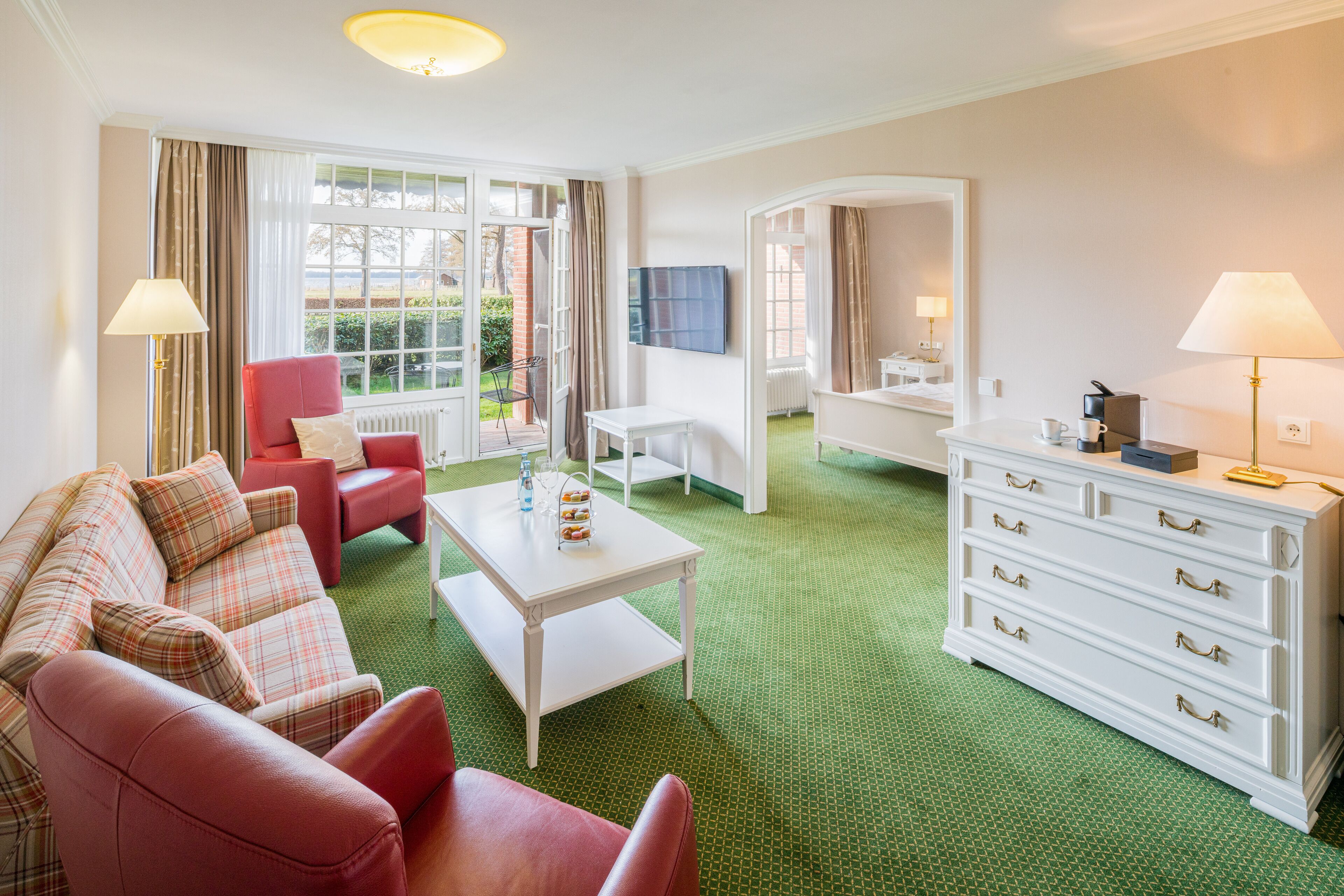 Una habitación de hotel con un sofá rojo, una mesita de café blanca, un tocador blanco y una alfombra verde.