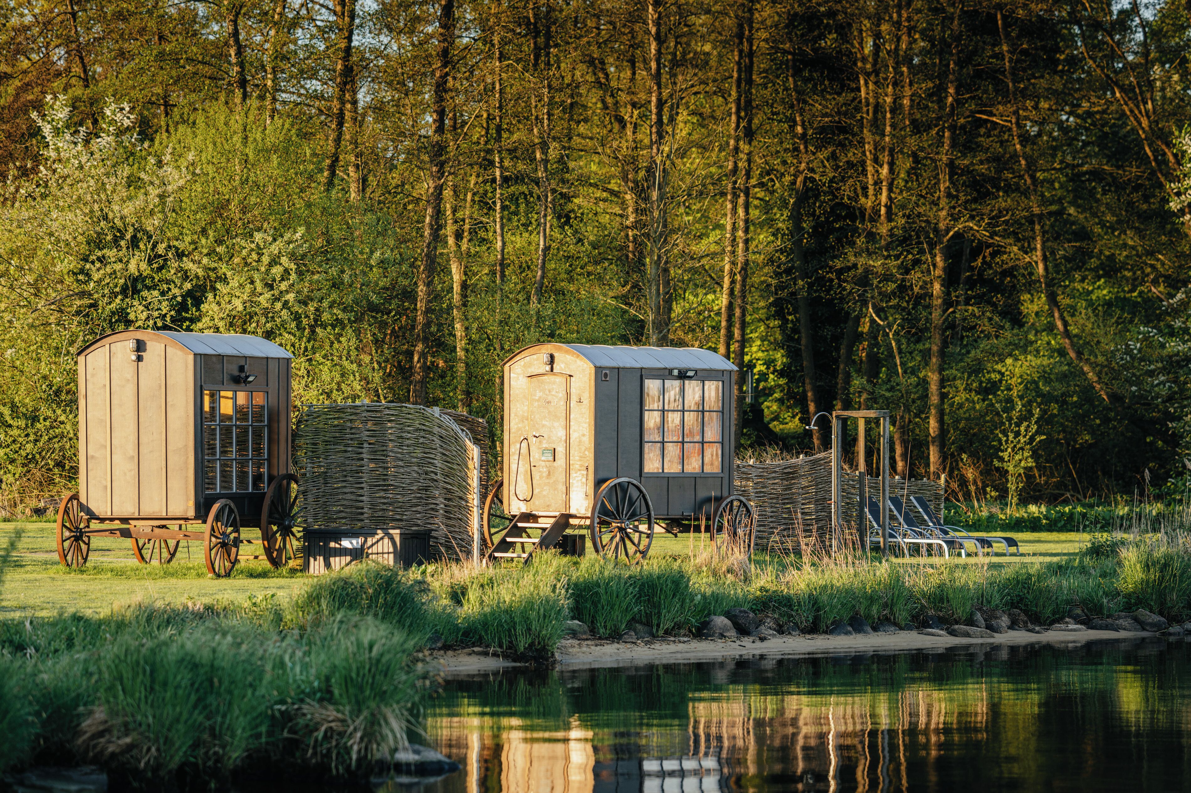 Two wooden cabins with attached wagons by a calm river, surrounded by dense forest.