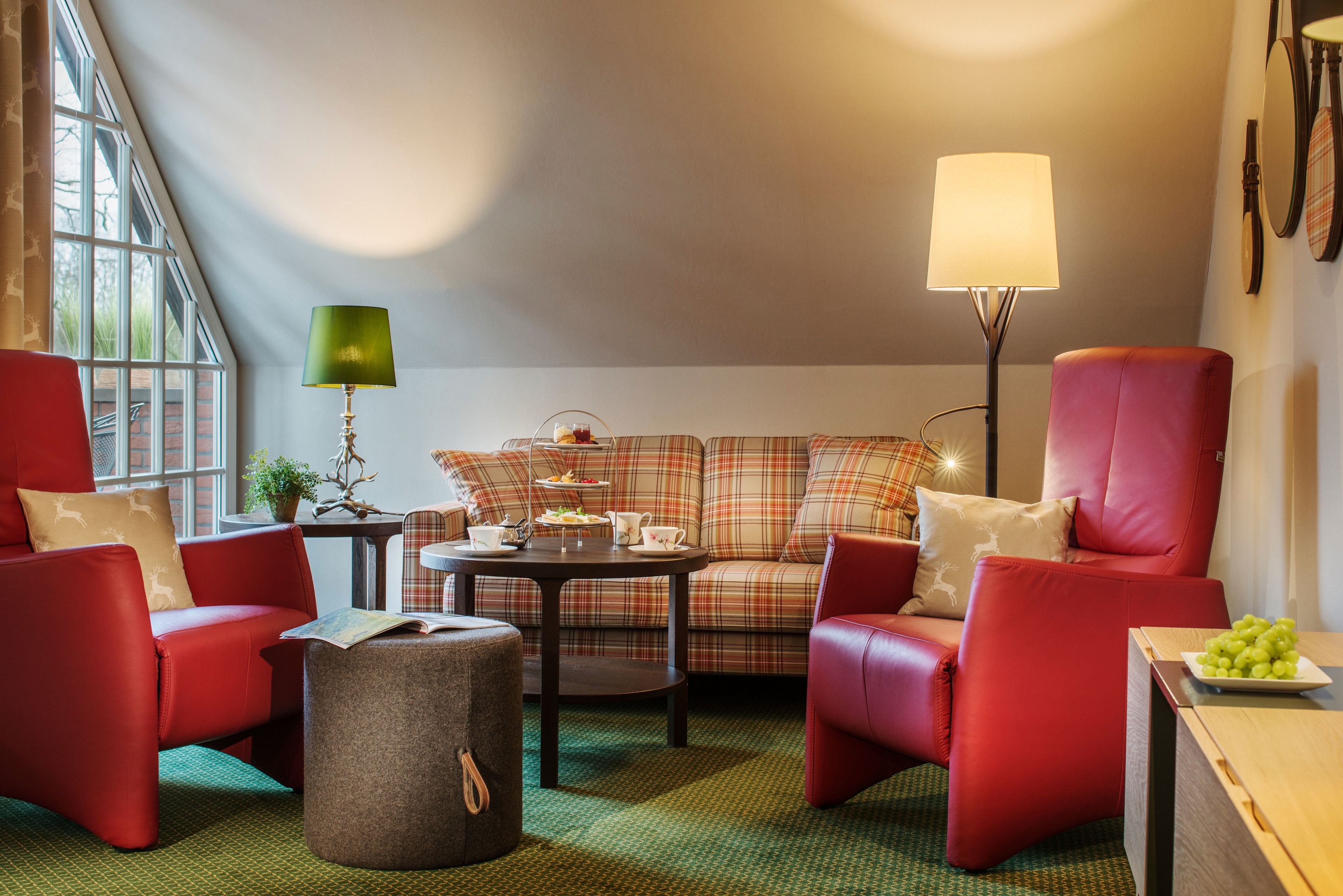 Een gezellige woonkamer met een geruite bank, rode stoelen, een tafeltje met een lamp en uitzicht op de omgeving door een groot raam.