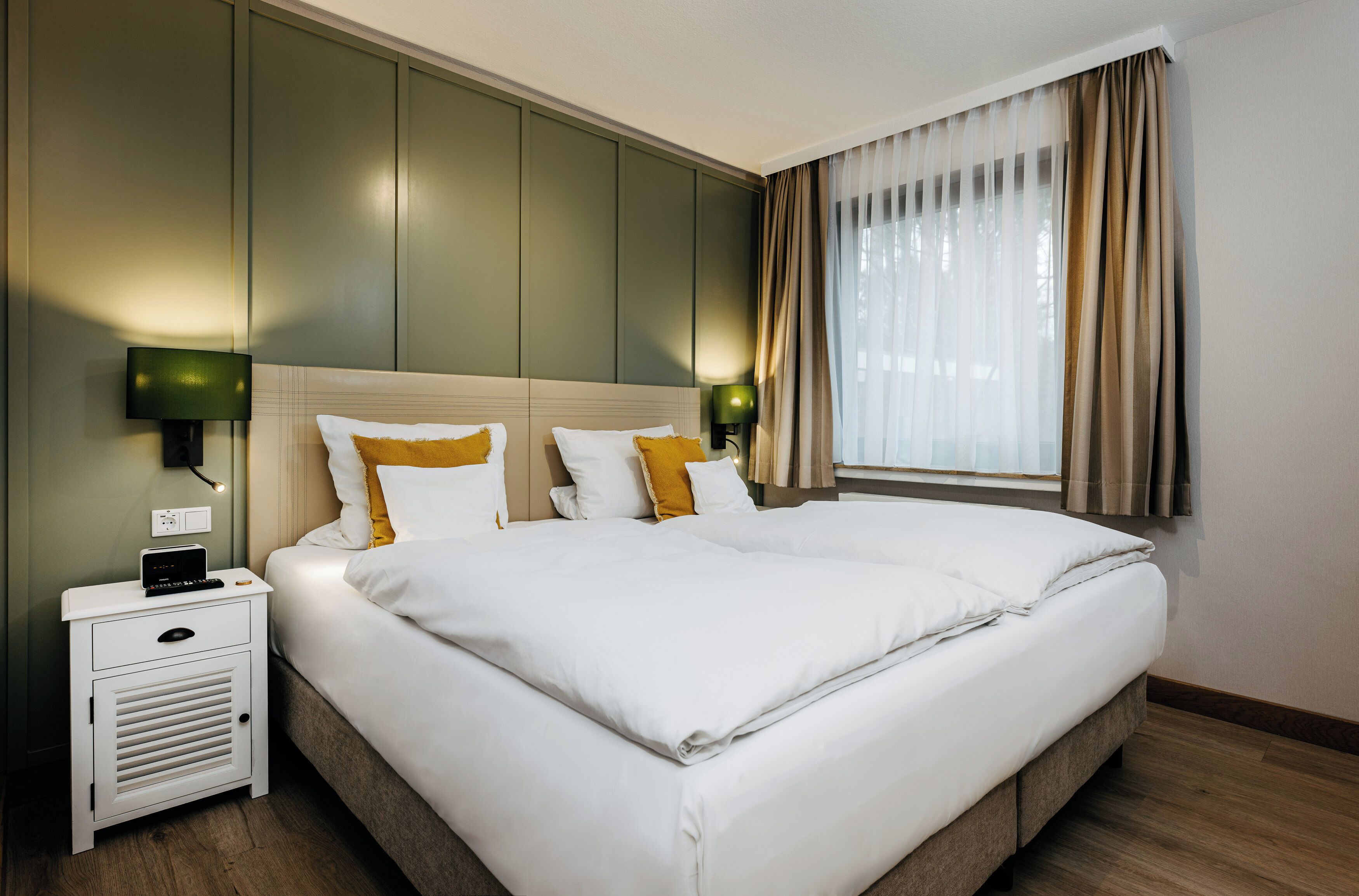A neatly made double bed with white linens and two pillows, a bedside table with a lamp, and a window with curtains.