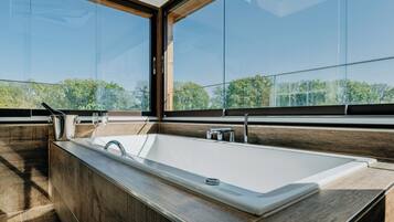 Un baño moderno con una bañera grande, piso de madera y ventanas amplias que dan a vista de árboles.