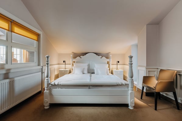 Un dormitorio ordenado con una cama grande, mesitas de noche, una silla y un ventanal con persianas.