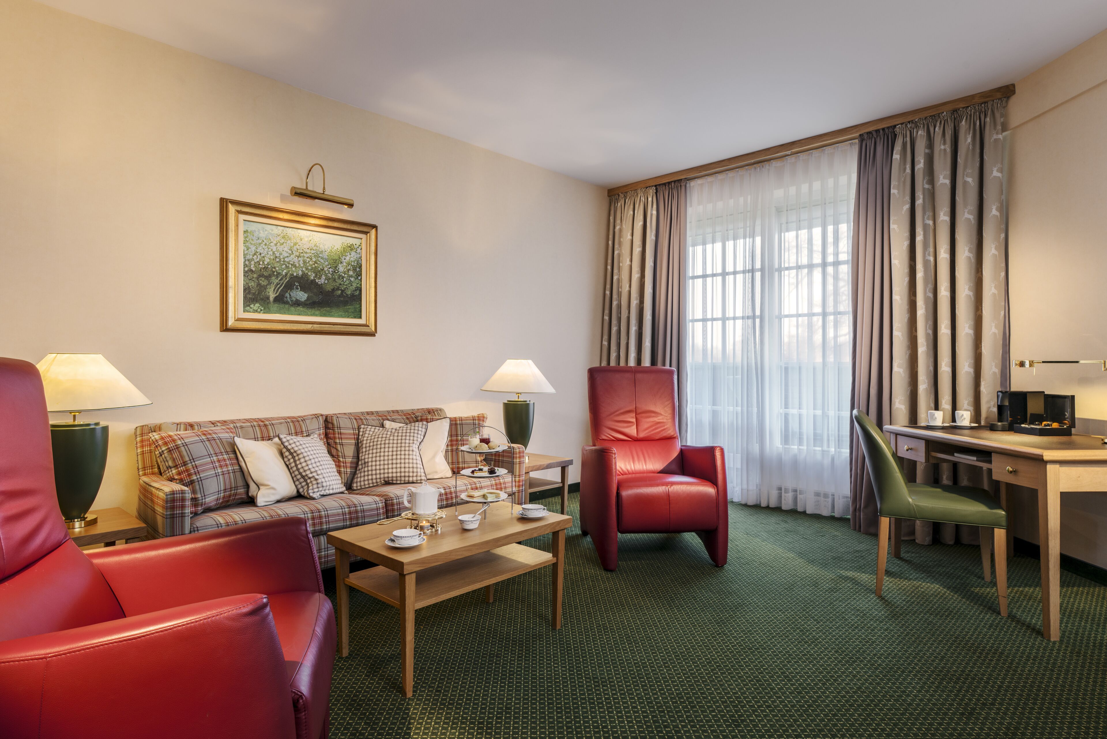 Ett hotellrum med en rutig soffa, en röd läderfåtölj, ett skrivbord i trä och en grön stol. Det finns en tavla på väggen och ett fönster med gardiner.