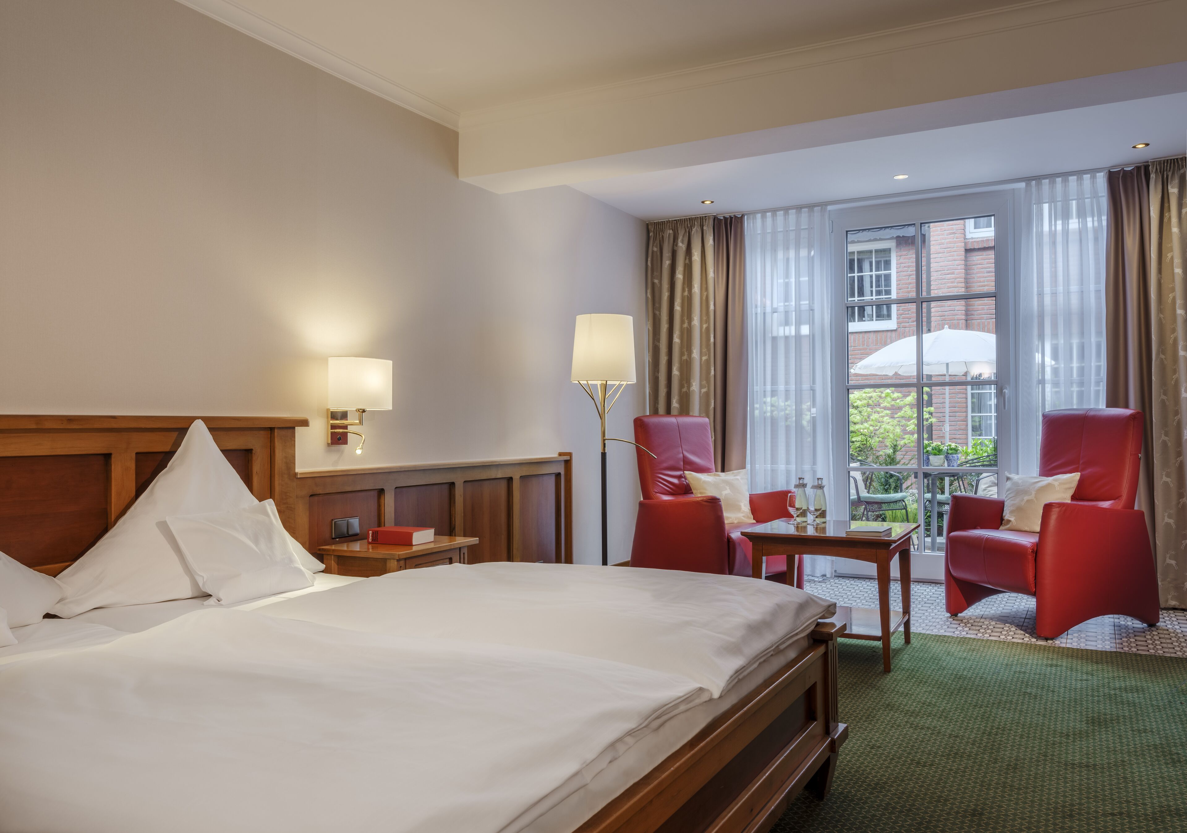 A hotel room with a large bed, two red chairs, a small table, and a window with curtains.