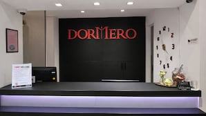 A reception desk with a black backdrop and the name 'DORMIREO' in red letters. There is a 'FANCY WELCOME' sign, a clock, and a jar of sweets on the counter. - DORMERO Hotel Coburg (Coburg)
