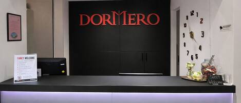 Una recepción con un fondo negro y el nombre 'DORMIREO' en letras rojas. Hay un cartel que dice 'FANCY WELCOME', un reloj y un tarro de caramelos en la encimera.