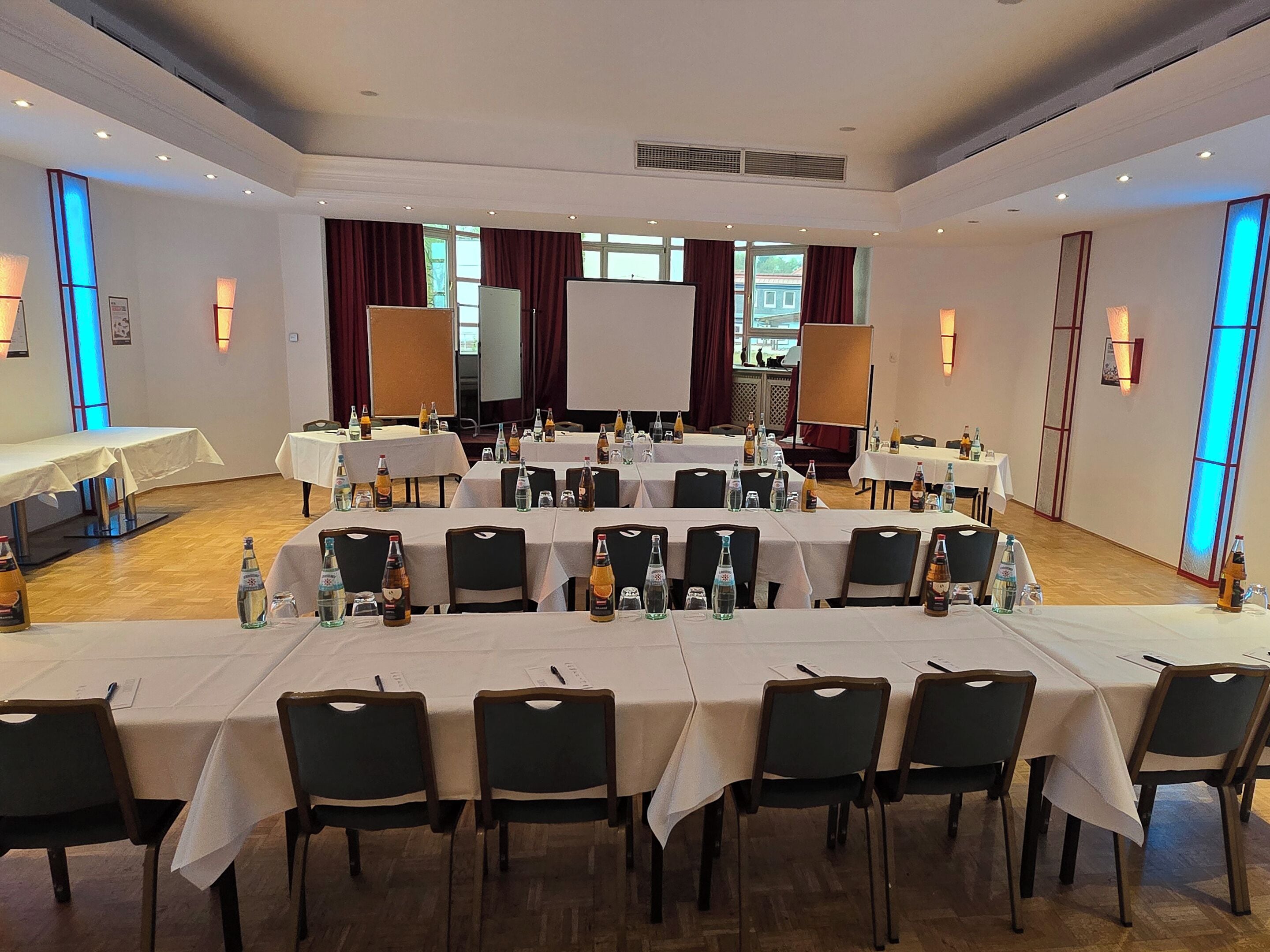 A conference room with multiple tables and chairs, equipped with water bottles and glasses, and large windows with curtains.