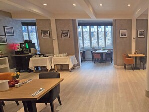 A spacious dining area with multiple tables and chairs, a counter with a coffee machine, and framed pictures on the walls. - DORMERO Hotel Coburg (Coburg)