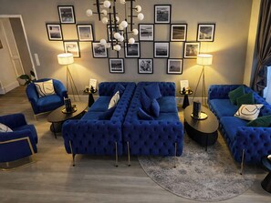 A modern living room with a blue sectional sofa, two armchairs, and a coffee table. The room features a large wall-mounted picture frame with multiple photographs, two floor lamps, and a patterned rug. - DORMERO Hotel Coburg (Coburg)