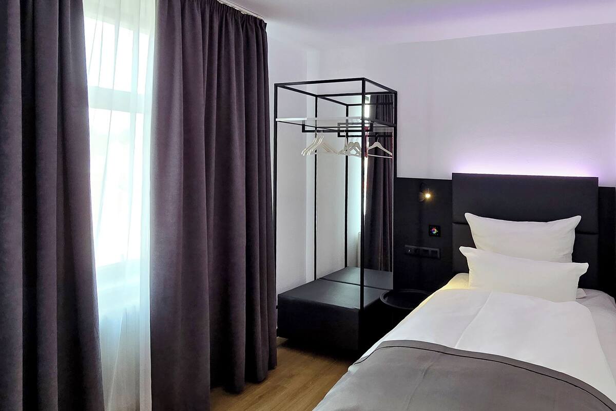 A modern hotel room with a bed, a black headboard, a bedside table, a mirror, and a clothing rack.