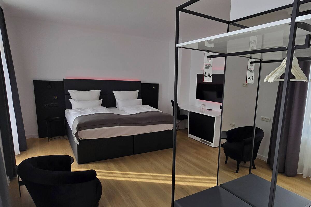 A modern hotel room with a large bed, a desk, a chair, and a wardrobe.