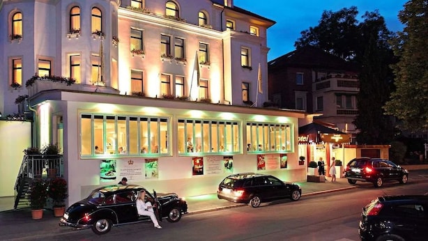 A hotel with a classic European architectural style, featuring a prominent entrance with a glass facade and a balcony with potted plants. - DORMERO Hotel Coburg (Coburg)