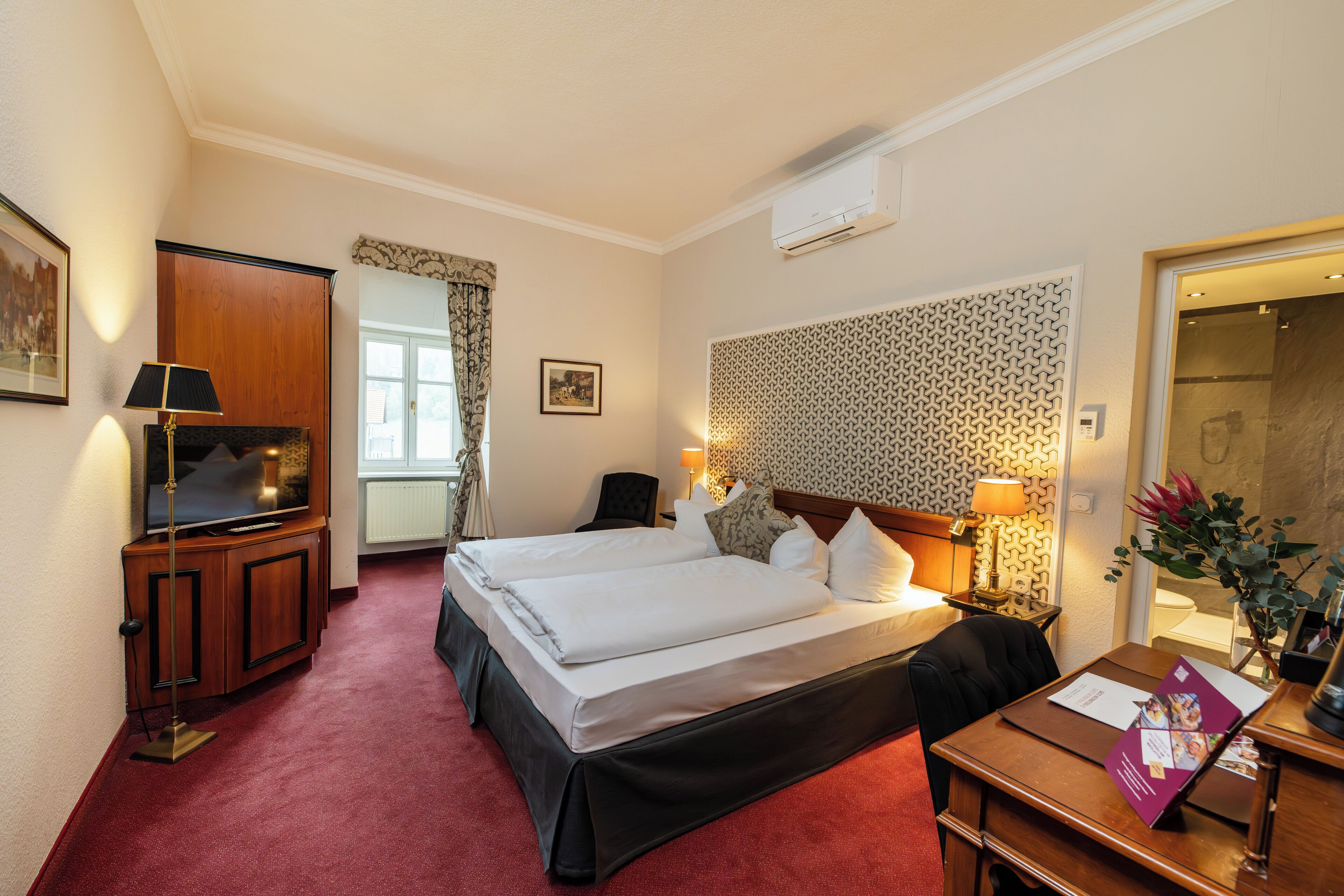 Une chambre d’hôtel avec deux lits, un bureau avec une chaise, une télévision, une lampe de bureau, une fenêtre avec des rideaux et un mur décoré de motifs.