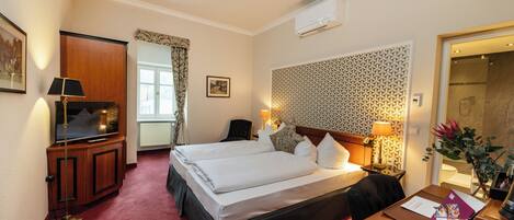 Une chambre d’hôtel avec deux lits, un bureau avec une chaise, une télévision, une lampe de bureau, une fenêtre avec des rideaux et un mur décoré de motifs.