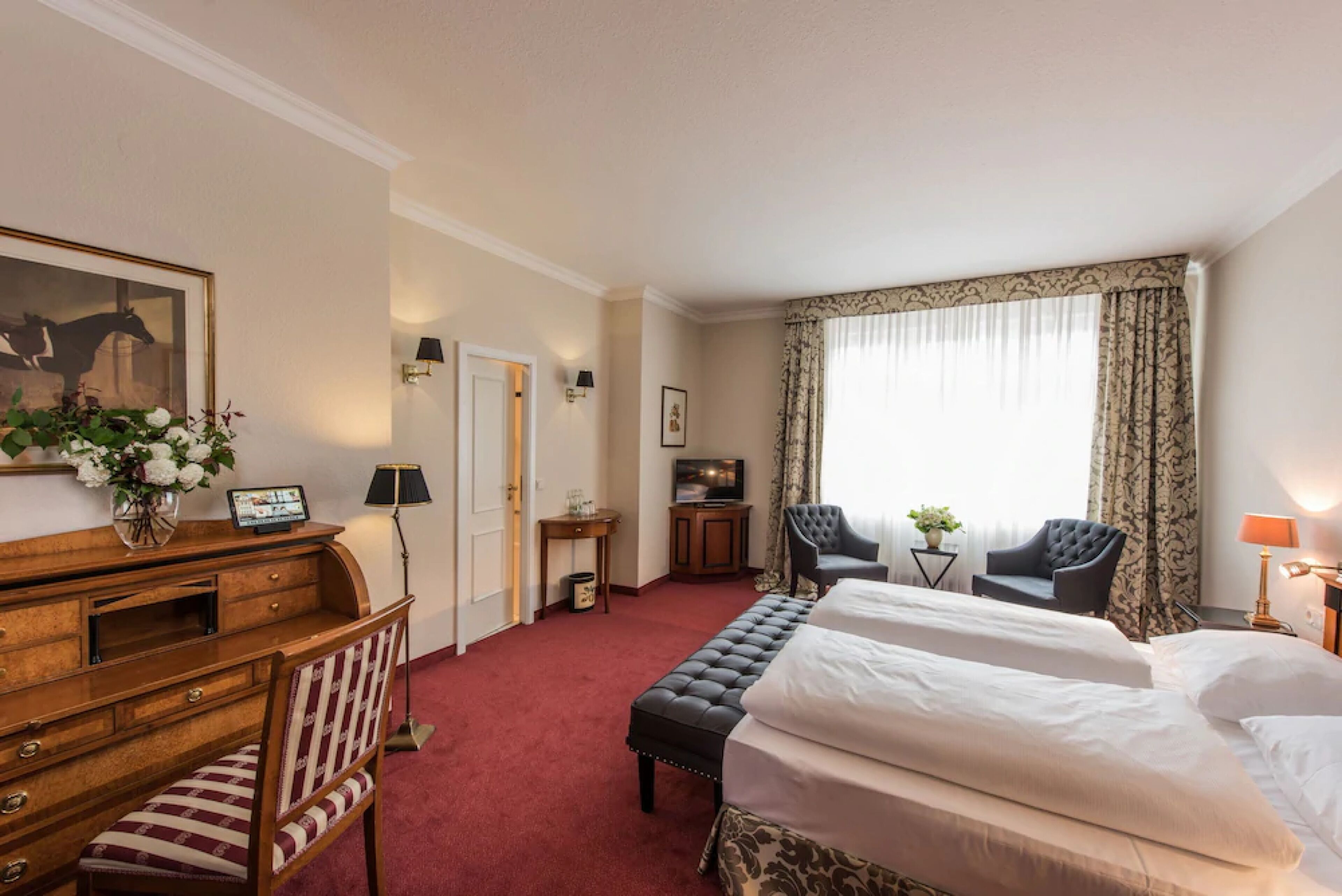 Habitación de hotel con dos camas, un escritorio con silla, televisión y un ventanal con cortinas.