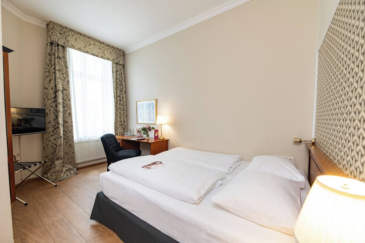 Hotellihuone, jossa on sänky, työpöytä, tuoli, lamppu ja ikkuna, jossa on verhot.