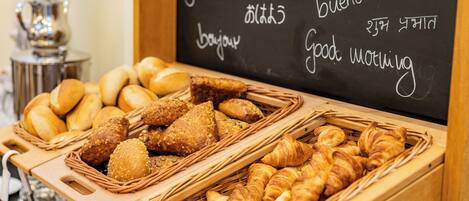 Un bufé de desayuno con panes y bollería variados, y un cartel con saludos en varios idiomas.