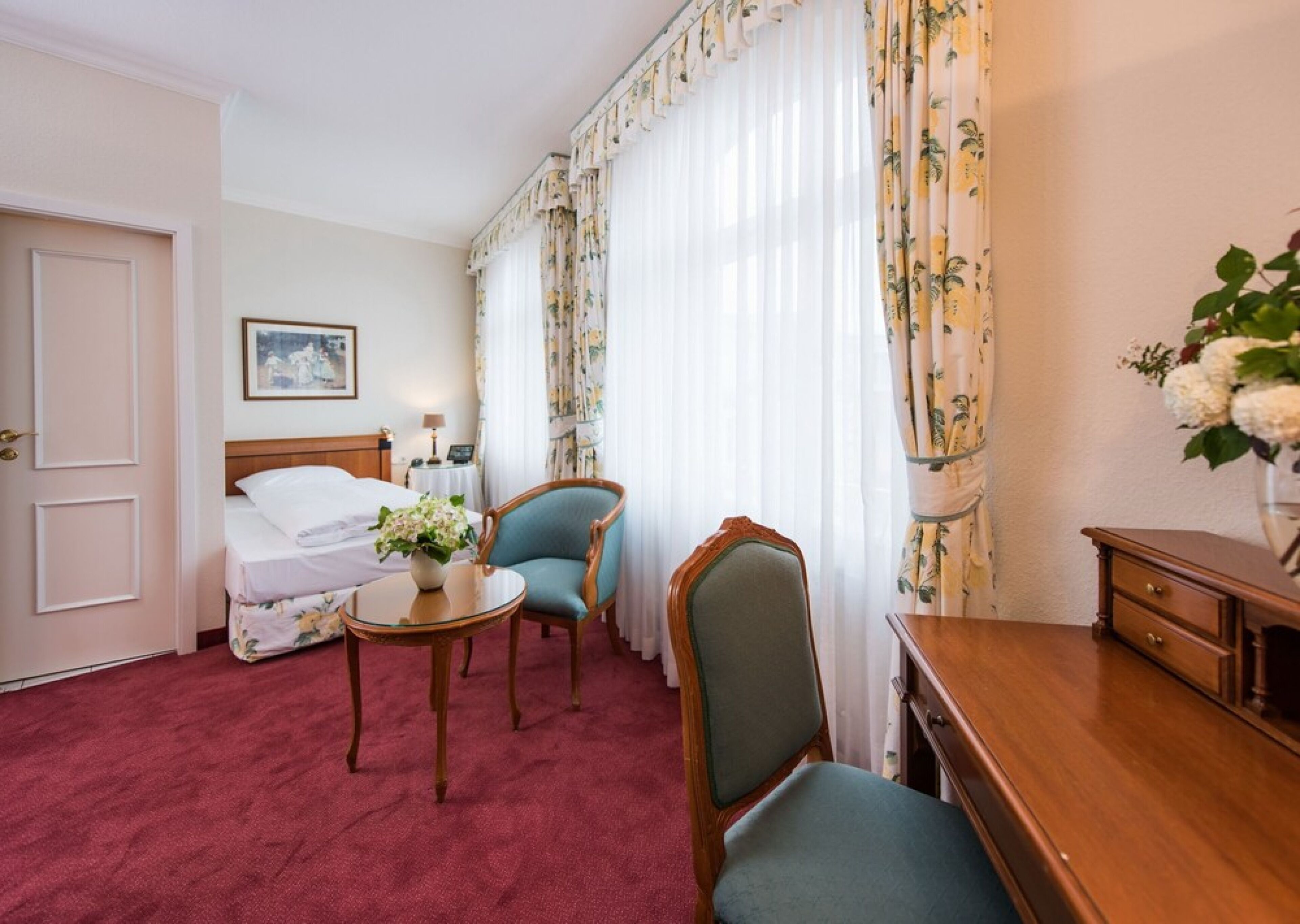 Habitación de hotel con cama, dos sillas, una mesita, un escritorio y un florero con flores.