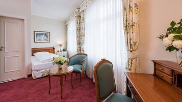 A hotel room with a bed, two chairs, a small table, a desk, and a vase with flowers.