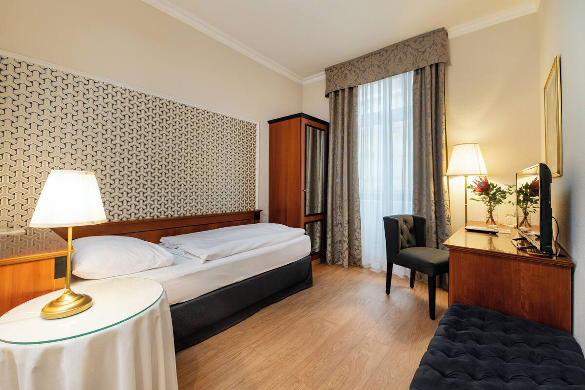Hotellihuone, jossa on sänky, yöpöytä lampun kanssa, työpöytä tuolilla, peili ja suuri ikkuna, jossa on verhot.