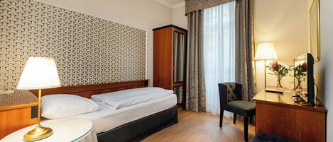 A hotel room with a bed, bedside table with a lamp, a desk with a chair, a mirror, and a large window with curtains.
