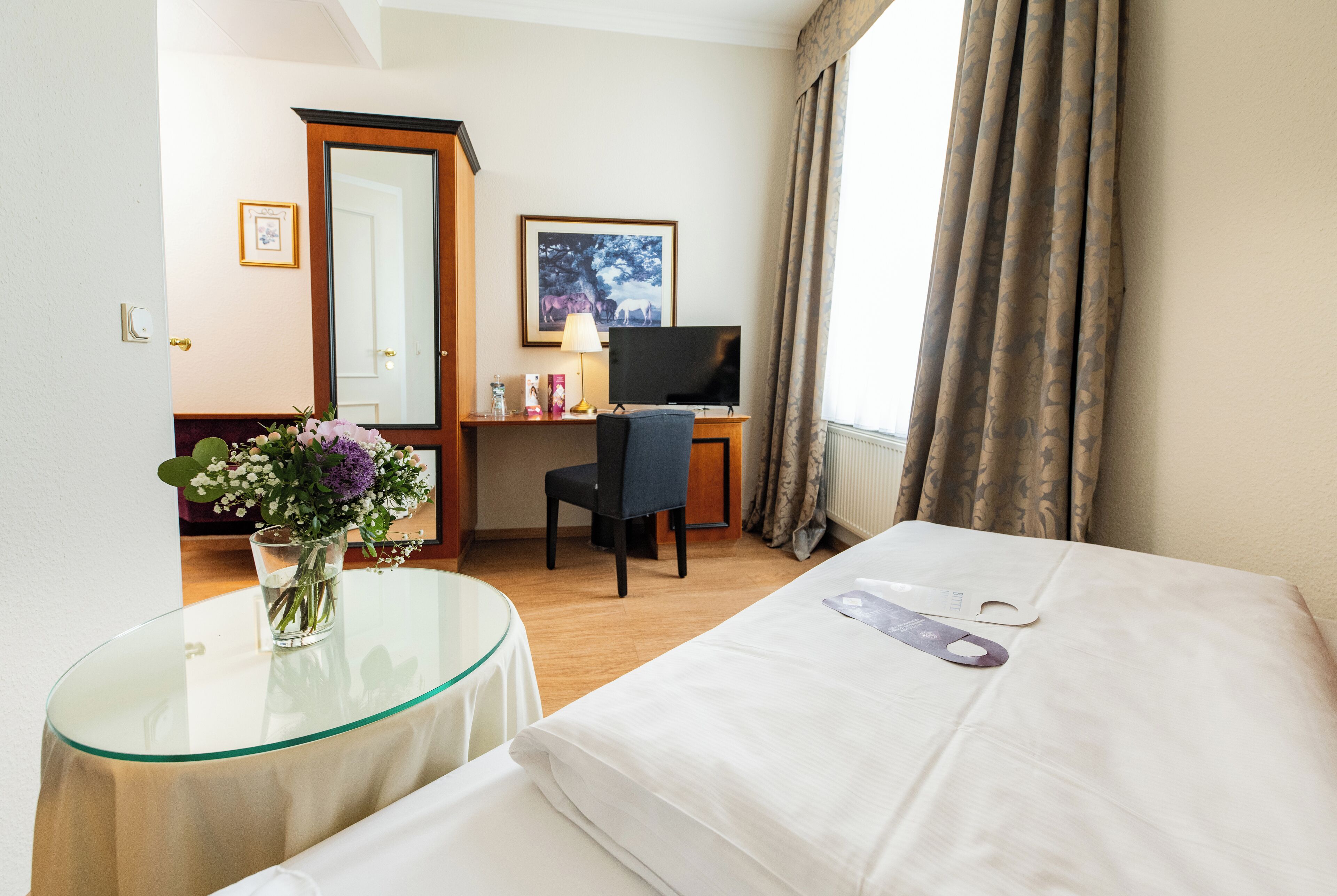 Habitación de hotel con cama, escritorio con computadora, silla, mesa de vidrio con un florero y ventana con cortinas.