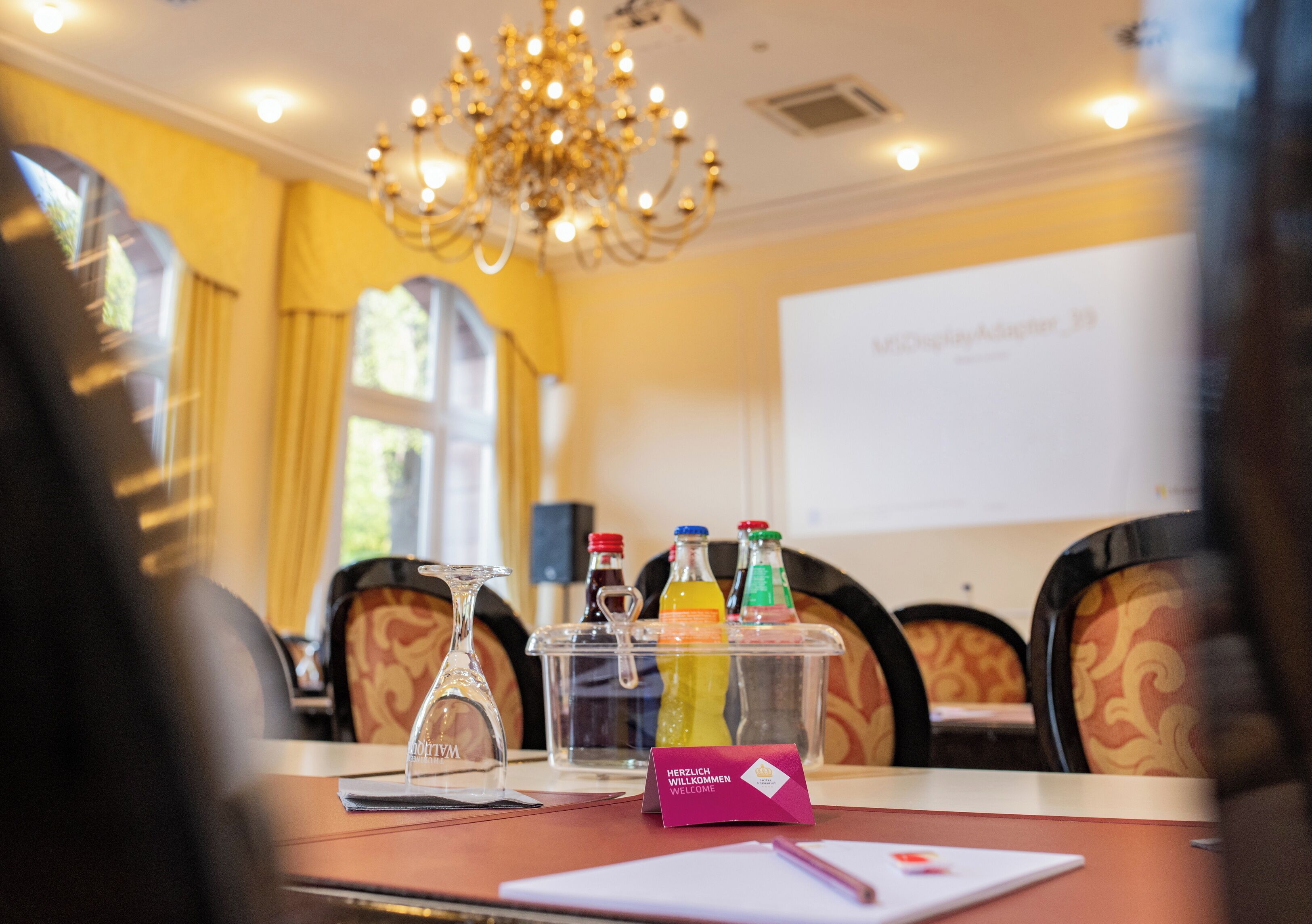 Un salón de conferencias con una pantalla de proyector, un decantador de vidrio y un porta vasos con bebidas.