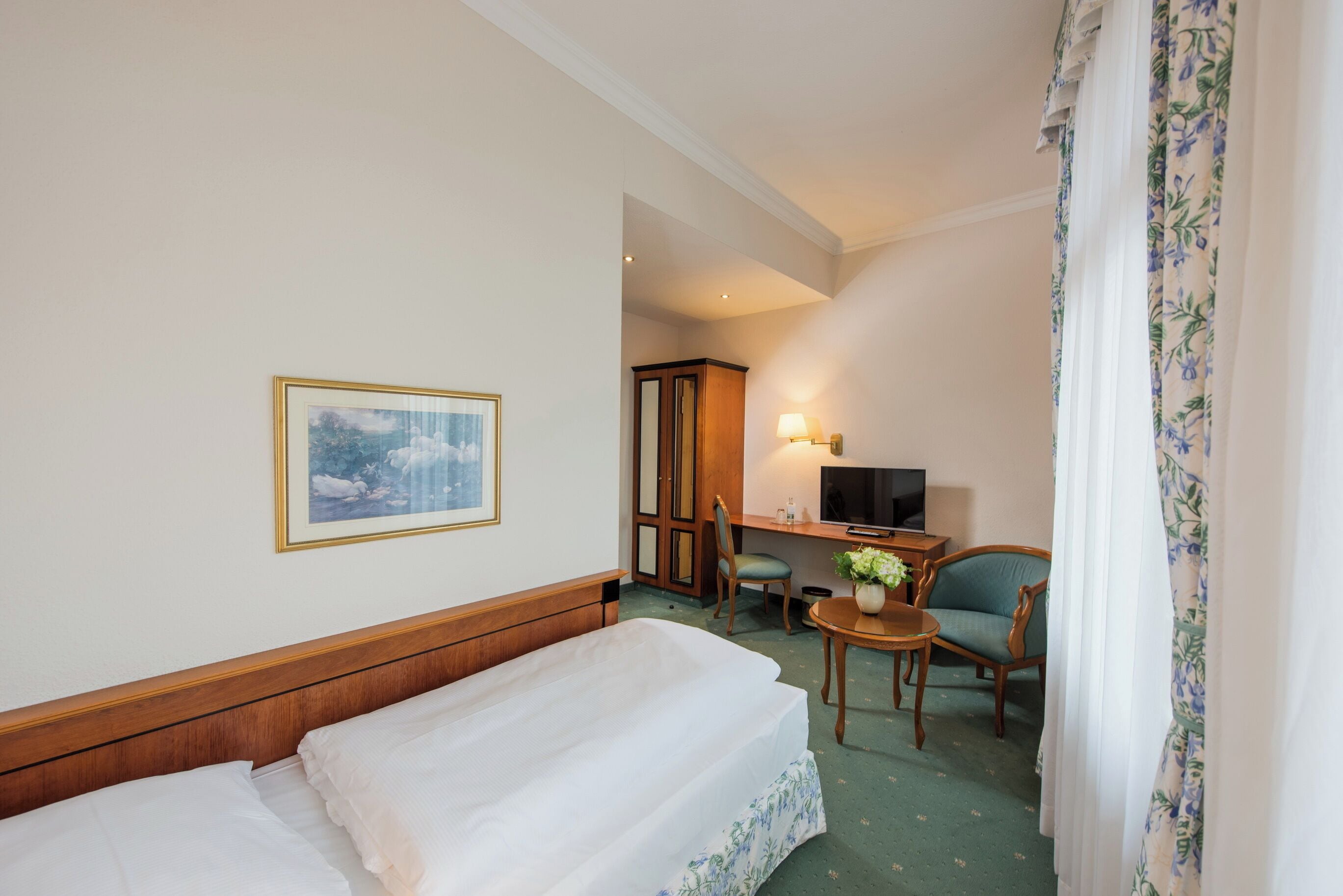 Quarto de hotel com uma cama, uma escrivaninha com cadeira, uma televisão e um quadro na parede.