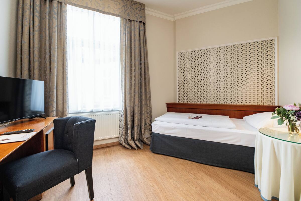 Quarto de hotel com cama, escrivaninha com cadeira, televisão e janela com cortinas.