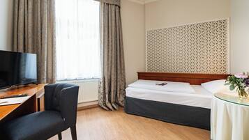 Quarto de hotel com cama, escrivaninha com cadeira, televisão e janela com cortinas.