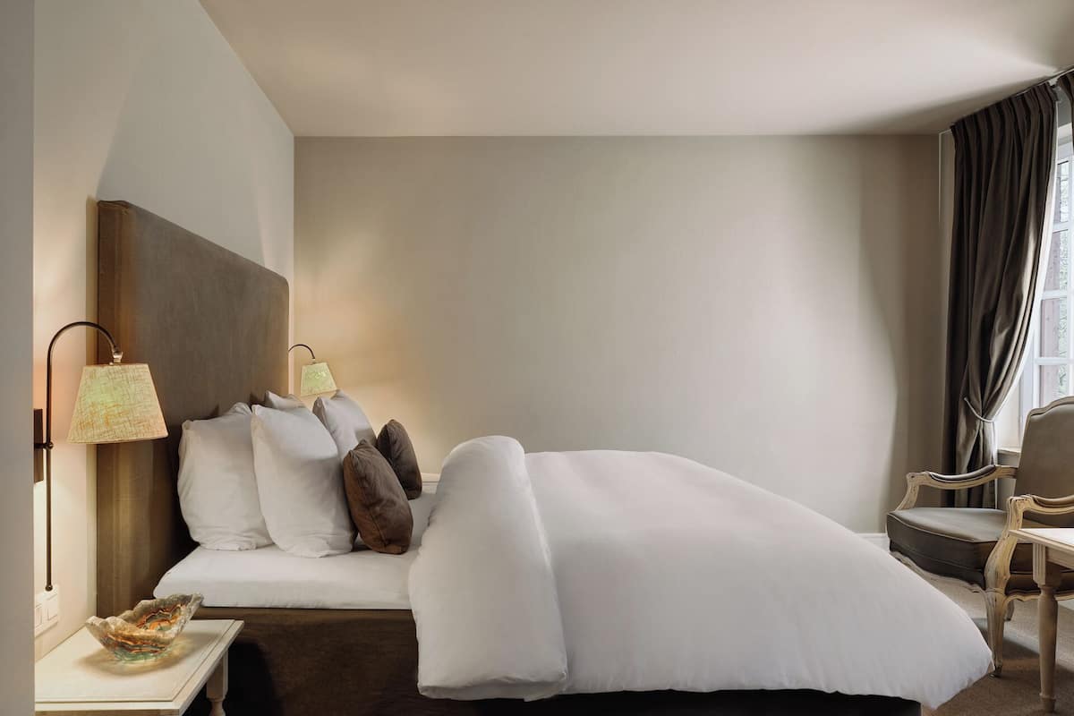Habitación de hotel con cama, mesita de noche, lámpara, silla y ventana con cortinas.