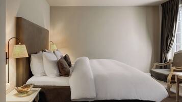 Habitación de hotel con cama, mesita de noche, lámpara, silla y ventana con cortinas.