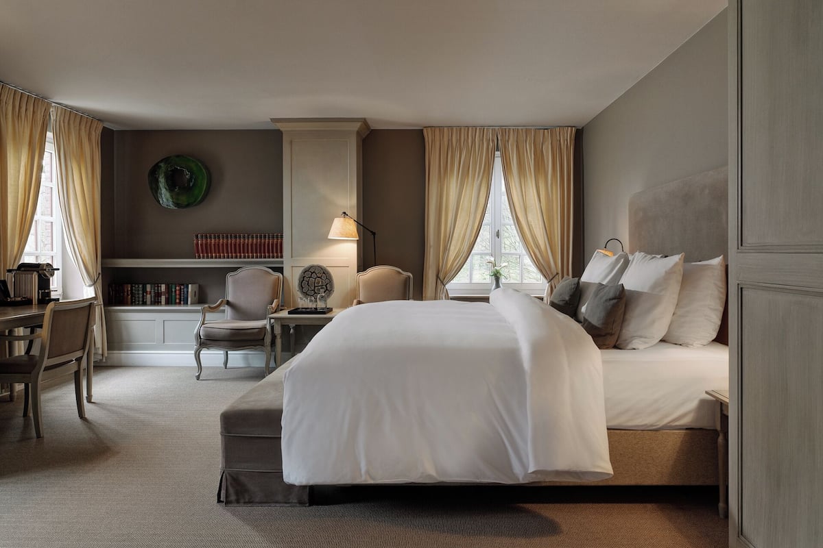 Habitación de hotel con una cama grande, un escritorio con lámpara, una silla, una estantería y una ventana con cortinas.