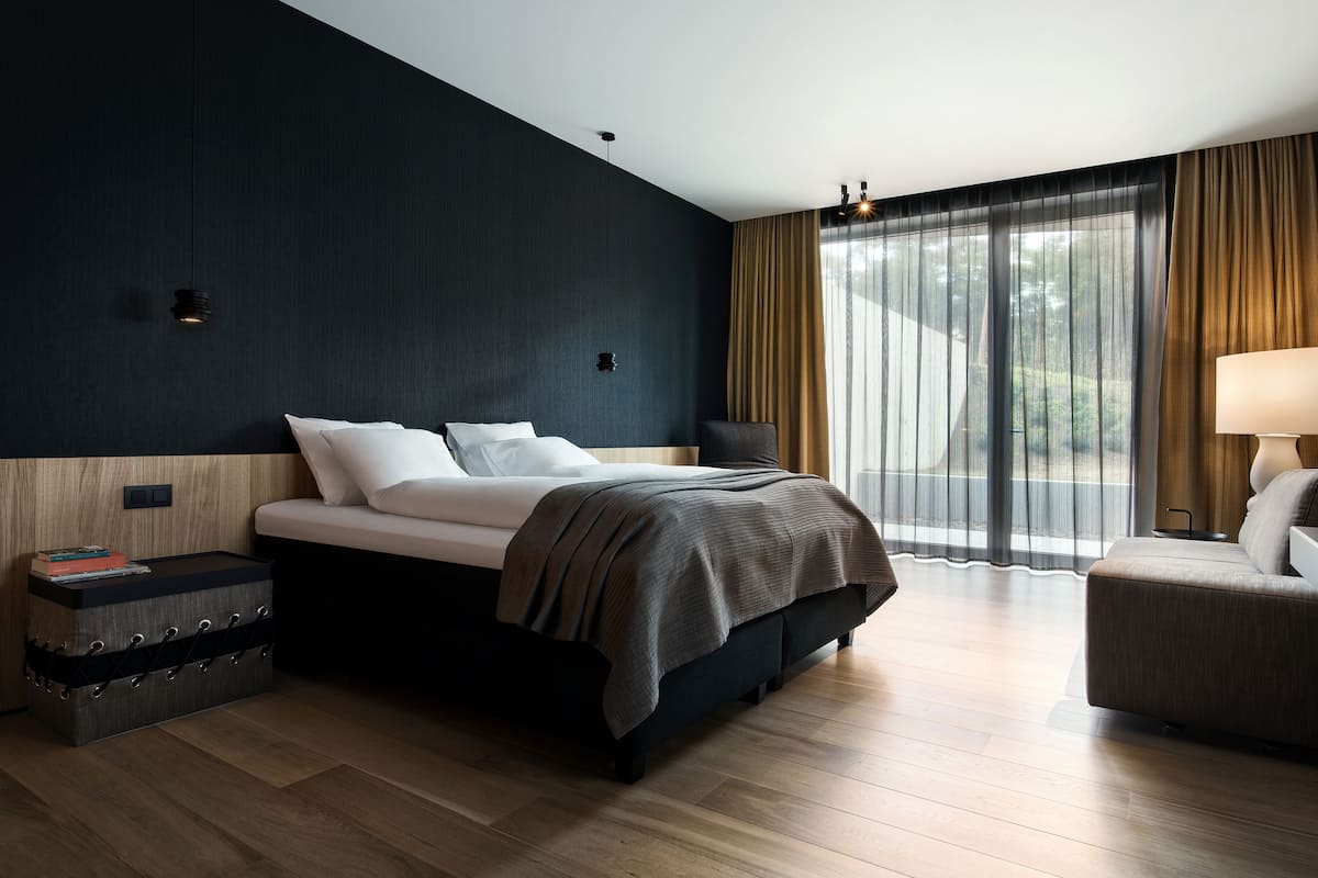 Una habitación de hotel moderna con una cama grande, una mesita de noche, una silla y vistas al exterior a través de cortinas translúcidas.