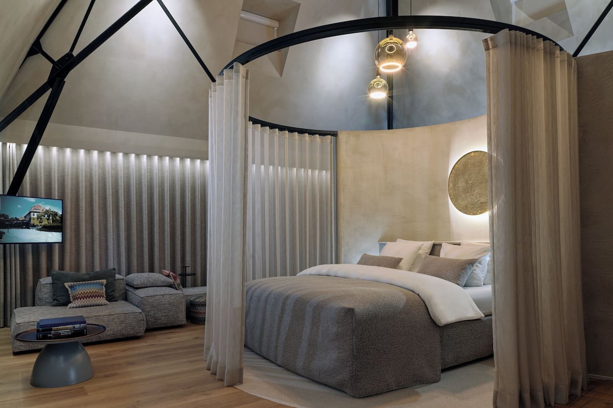 Un dormitorio moderno con techo curvo, cama con ropa de cama gris, televisor en la pared y un ventanal con cortinas.
