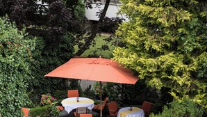 An outdoor patio area with round tables and orange chairs, surrounded by greenery and potted plants.