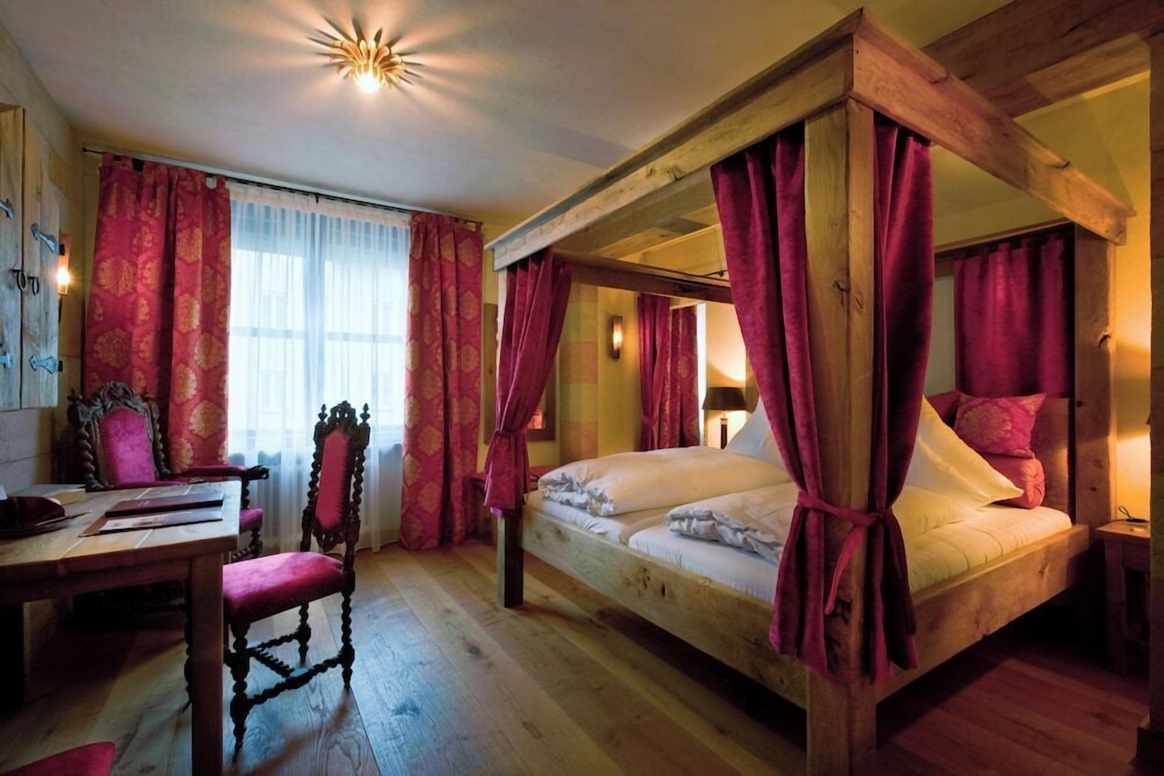 Ein Bett mit Baldachin, mit roten Vorhängen, ein Holztisch und ein Stuhl in einem Zimmer mit Holzböden.