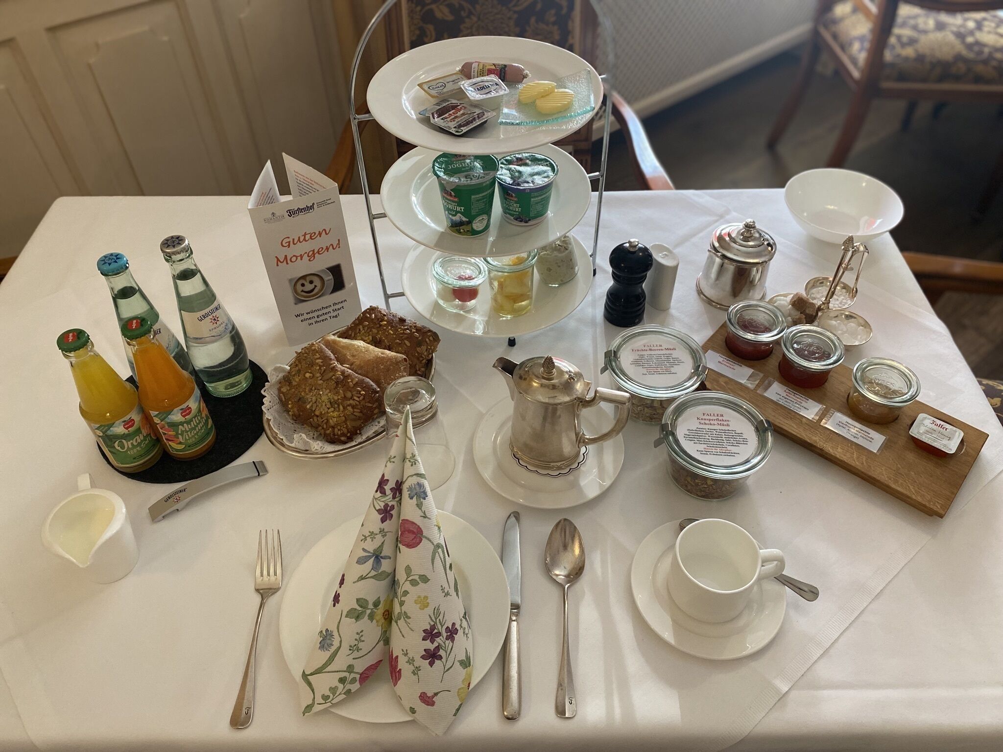 A breakfast setting with a three-tiered tea service, bottles of orange juice, a floral napkin, a silver teapot, and various condiments.