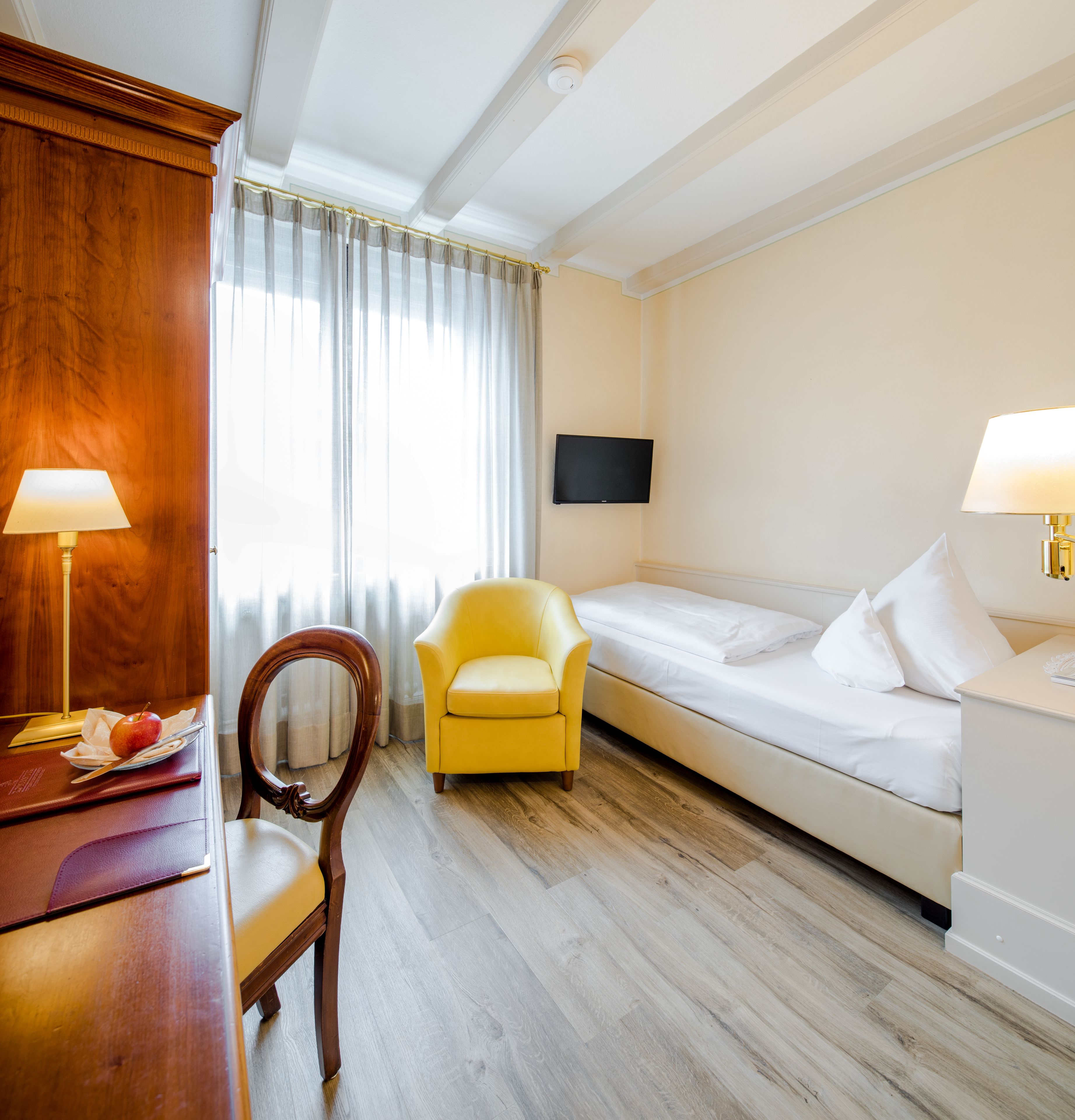 A hotel room with a wooden desk, a yellow armchair, a bed, and a flat-screen TV on the wall.