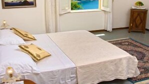 A bedroom with a bed, a window with a view of a mountain, a painting on the wall, and a small table with a lamp. - Villa Cheta Hotel & Restaurant (Maratea)