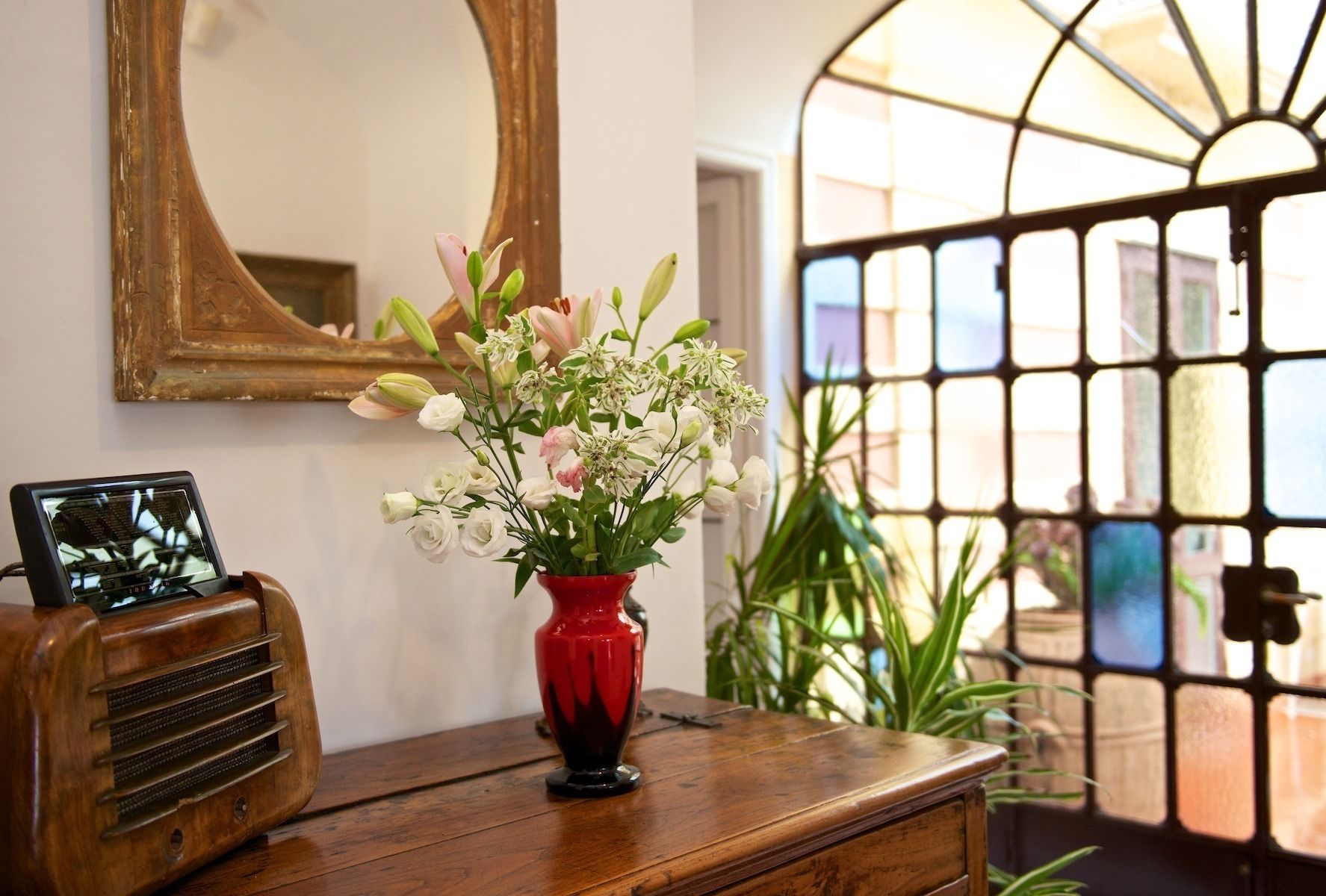 Une commode en bois, sur laquelle sont posés un vase rouge rempli de fleurs, une radio ancienne et un miroir accroché au mur.
