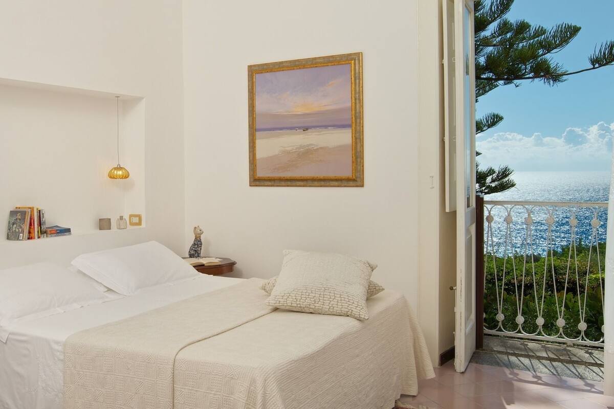 Une chambre à coucher avec un lit, des tables de chevet, une commode, un tableau encadré et une vue sur la mer depuis une porte-fenêtre donnant sur un balcon.