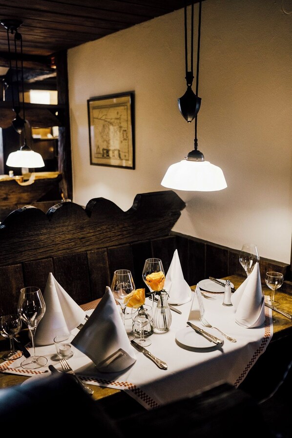 Mesa de comedor con manteles blancos, cubiertos y copas de vino, con una lámpara colgante arriba.