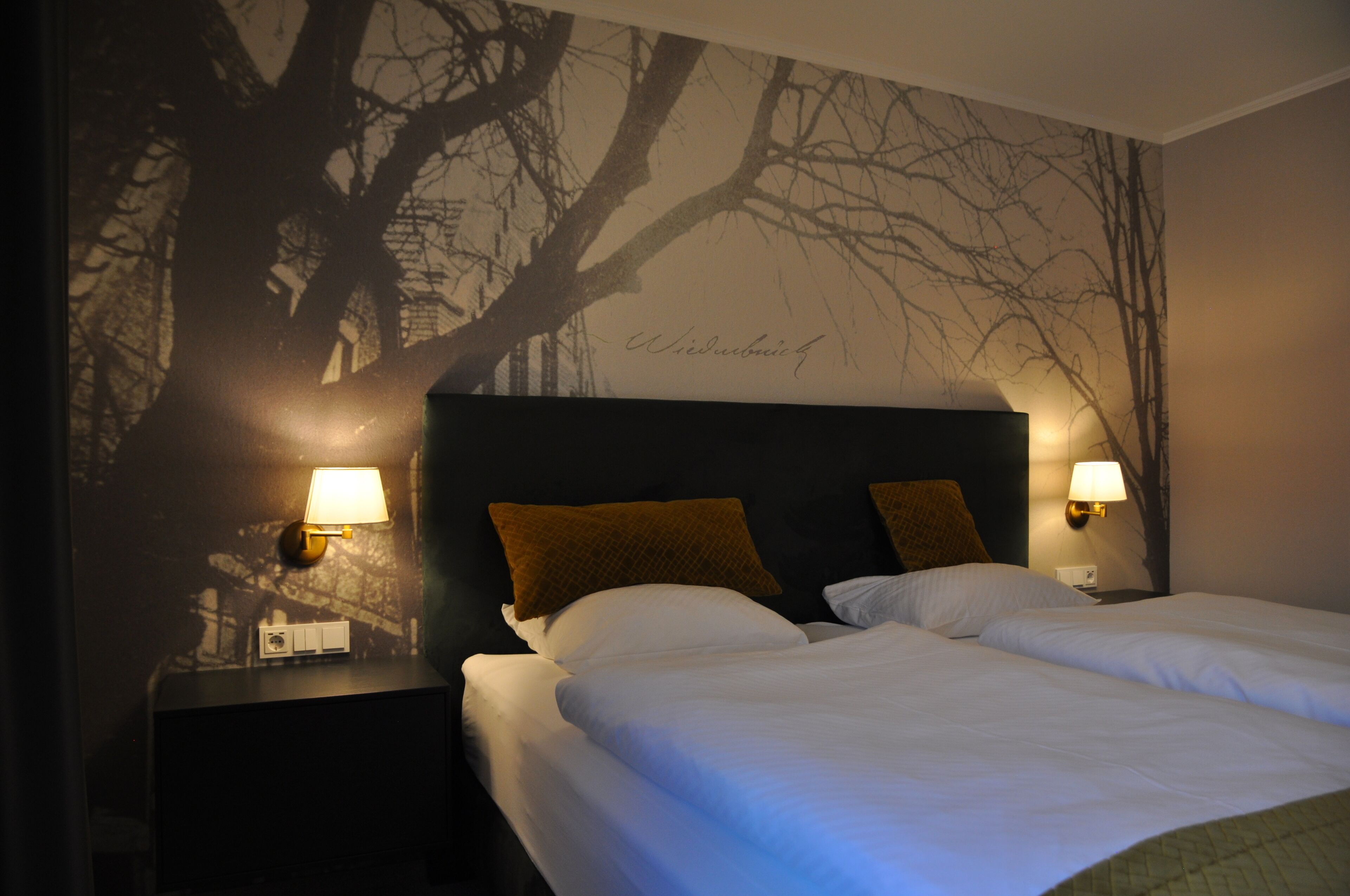 Habitación de hotel con una cama grande, dos lámparas de noche y un mural en la pared con árboles desnudos.