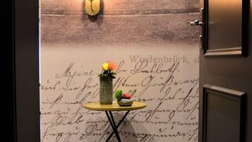 A room with a wall-mounted light fixture, a small table with a vase of flowers, and a door with a number sign.