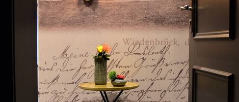 A room with a wall-mounted light fixture, a small table with a vase of flowers, and a door with a number sign.