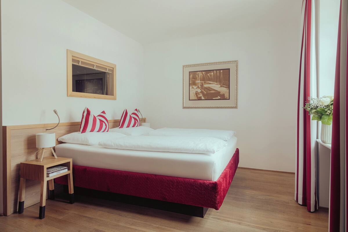 Habitación de hotel ordenada con cama, mesita de noche con lámpara, un cuadro en la pared y ventana con cortinas.