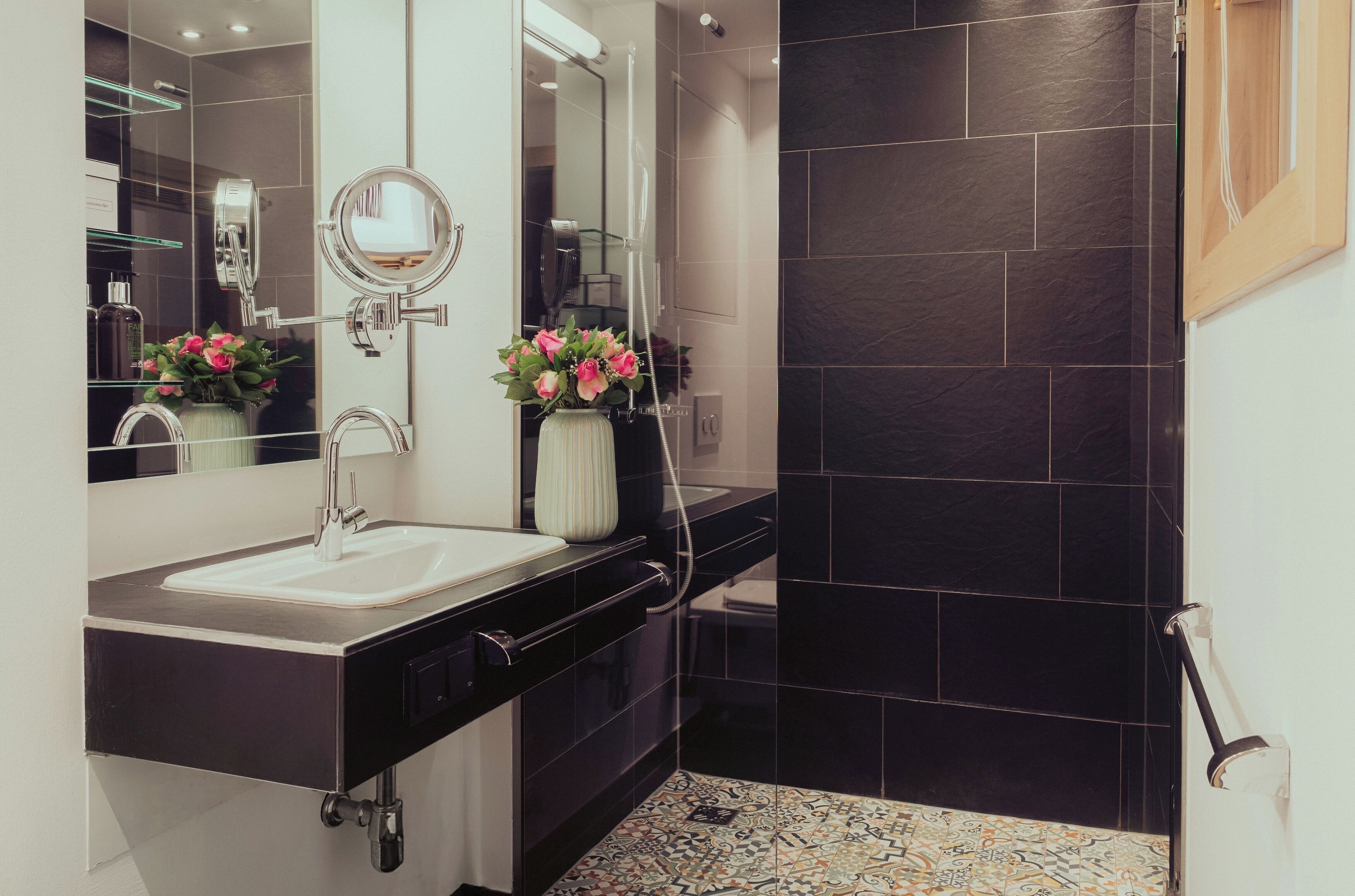 A modern bathroom with a black tiled shower, a white sink with a mirror, and a vase with pink flowers.